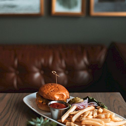 A plate with a burger, fries, and a dip sits on a table in a cozy setting with a leather couch and framed pictures in the background.