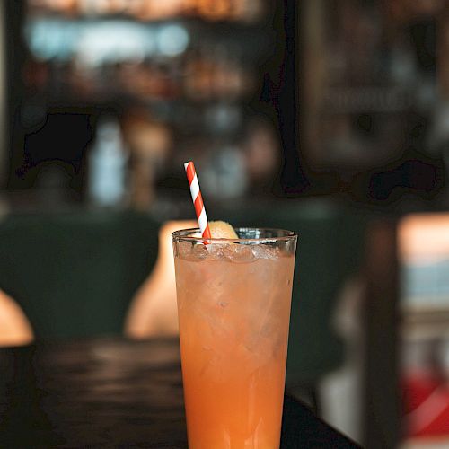 A tall glass filled with an orange beverage, ice cubes, and a striped straw sits on a dark table in a dimly lit bar.