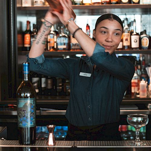 A bartender is shaking a cocktail shaker behind a bar stocked with various bottles, with a bottle of liquor and a glass on the counter in front.