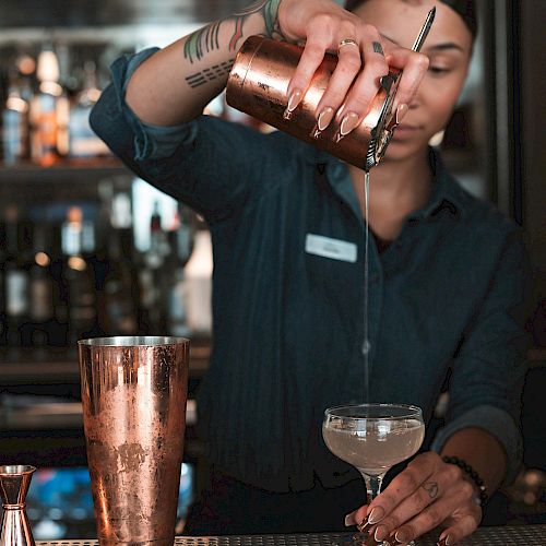 A bartender is pouring a cocktail from a shaker into a glass, with a backdrop of liquor bottles.