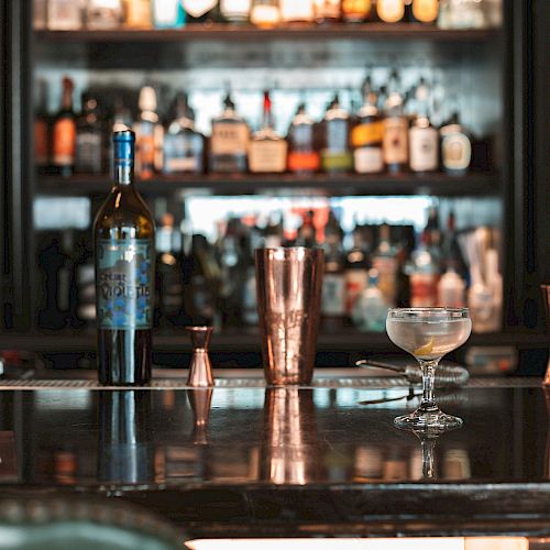 A bar counter with various bottles in the background, a wine bottle, cocktail shaker, and a glass with a drink on the counter.