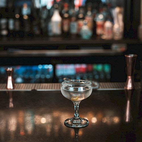 A cocktail glass sits on a bar counter, with shelves of various liquor bottles blurred in the background.