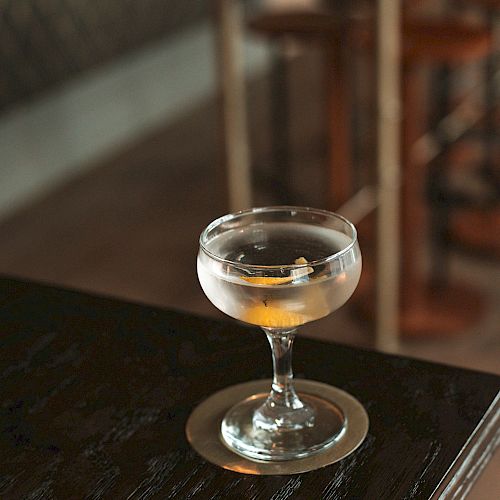 A cocktail in a coupe glass is placed on a wooden table, with a coaster beneath it. The background shows a wooden bar stool and a patterned wall.