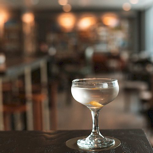 A single cocktail glass with a clear drink and a garnish sits on a dark wooden table in a blurred, warmly-lit bar setting.