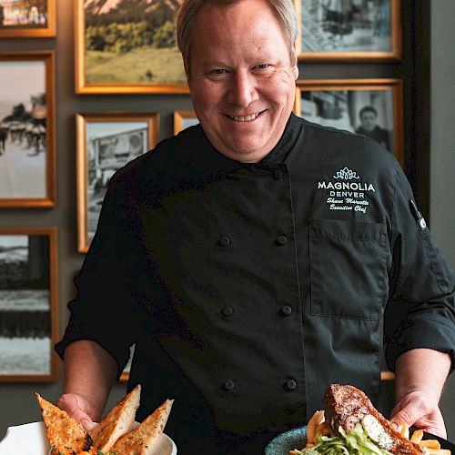 A smiling chef in a black uniform holds two plates of food in a restaurant, with framed photos behind him on the wall.