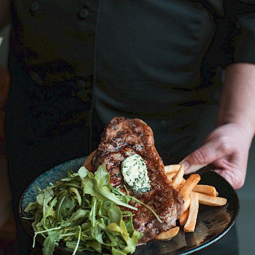 The image shows a person holding a plate with steak topped with a dollop of herb butter, French fries, and a green salad.