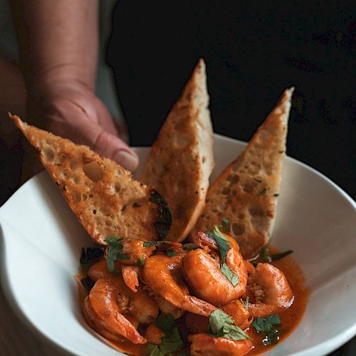 A person holding a white bowl filled with shrimp in sauce, garnished with parsley and accompanied by three pieces of toasted bread.