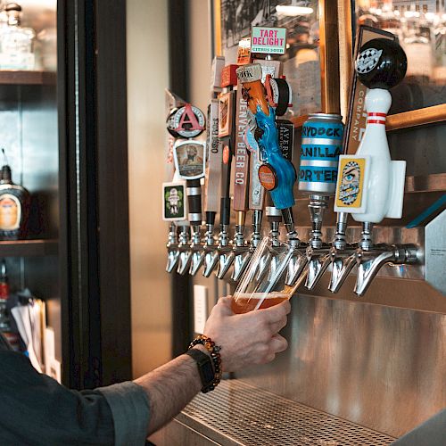 A person is pouring a drink from one of several beer taps at a bar, with colorful tap handles and framed pictures in the background.