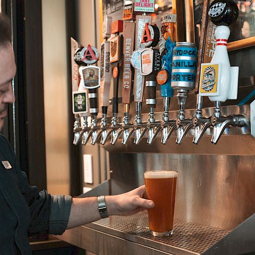 A person is pouring a beer from a tap at a bar with multiple colorful beer taps in the background, featuring various brewery logos and artwork.