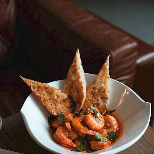 A dish featuring shrimp in a flavorful sauce, garnished with three toasted bread slices, presented on a wooden table next to a leather couch.