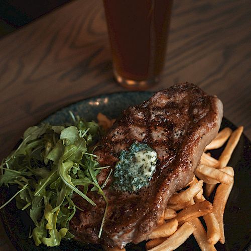 The image shows a plate with a grilled steak, fries, salad, and a dollop of herb butter, accompanied by a glass of beer on a wooden table.