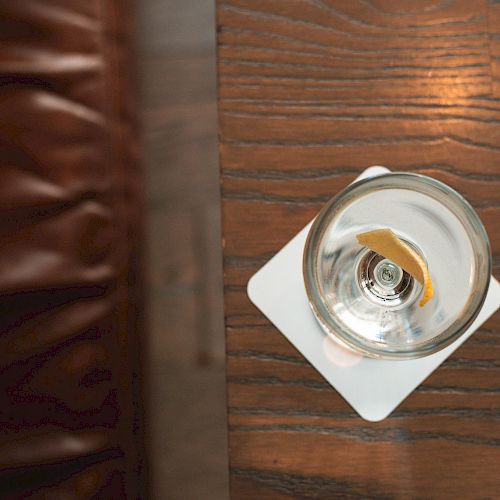 A glass with a drink and a citrus twist on a white coaster is placed on a wooden table next to a brown leather cushion, viewed from above.