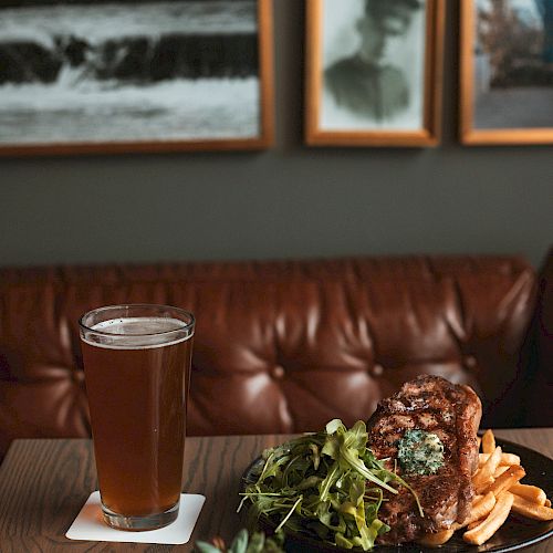 A meal with steak, fries, and greens is on a table beside a glass of beer. In the background, there are framed pictures on a wall.