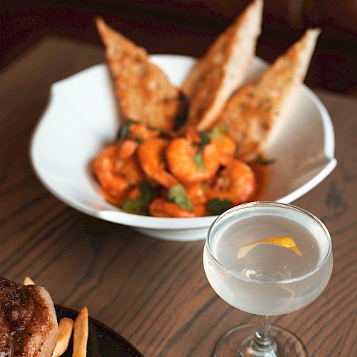 A plate of shrimp with bread slices, a glass of drink with a lemon slice, and a partial view of a steak and fries on a wooden table.