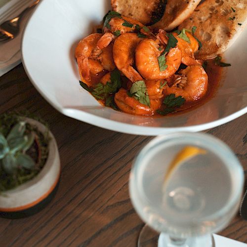 A plate of shrimp with sauce and bread, accompanied by a succulent plant, cocktail, and a napkin with 