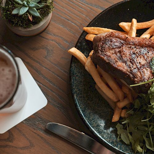 A plate with steak, fries, and greens alongside a beer on a coaster and a succulent plant on a wooden table.