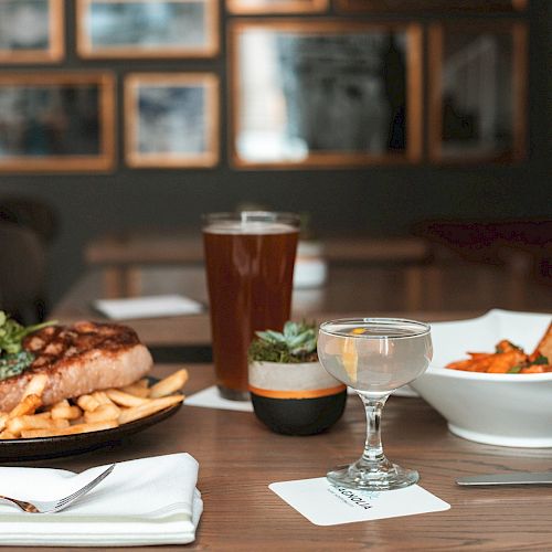 The image shows a table set with a plate of steak and fries, a bowl of soup with bread, a glass of beer, and a cocktail glass.