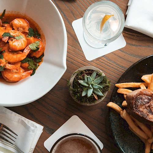 A table with shrimp dish, steak with fries, bread slices, a drink with lemon slice, and utensils on a wooden surface, centered by a small plant.