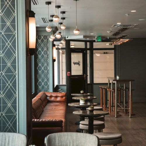 An indoor lounge/bar with a brown leather sofa, round side tables and stools, and hanging globe lights under a modern ceiling.