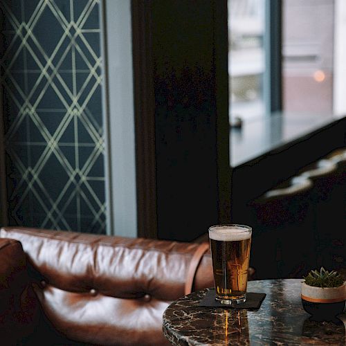 A cozy lounge with a leather sofa, marble table, a glass of beer, and a small plant, set beside a window with a geometric patterned wall.