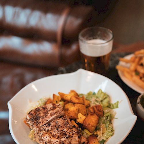 A plate with grilled meat and vegetables next to a glass of beer and a bowl of fries, placed on a table near a leather couch.