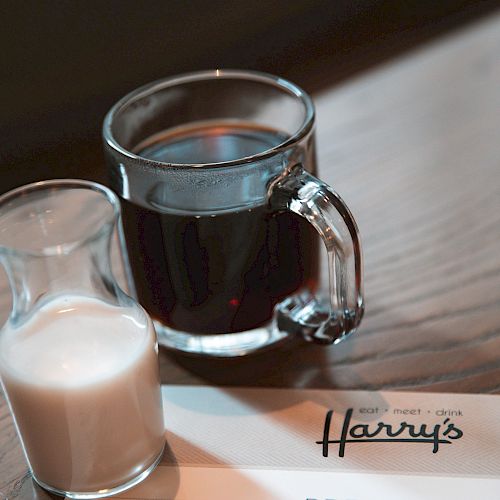 A glass of coffee, a small glass jug of milk, and a breakfast menu from a place called Harry's are on a wooden table.