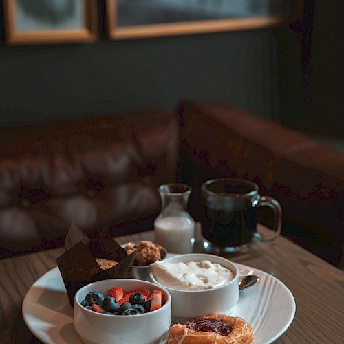 A plate with yogurt, berries, a pastry, and muffins. There's a small pitcher, a mug on the table, a leather sofa, and framed art in the background.