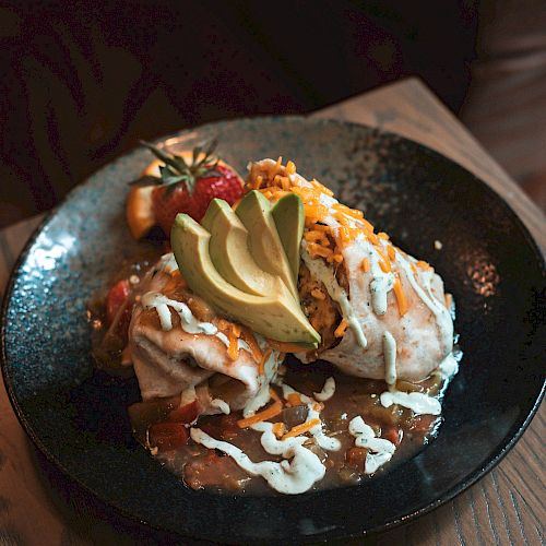 A black plate of food with a serving of meat or fried food topped with avocado slices, white sauce, strawberries, and herbs.