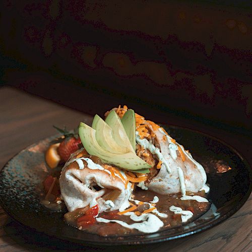 A dish garnished with creamy white sauce, avocado slices, and other ingredients on a black plate, placed on a wooden table.