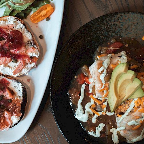 The image shows a plate with two pieces of bread with cream cheese, salmon, and onions, next to a dish with a loaded burrito topped with avocado and cheese.