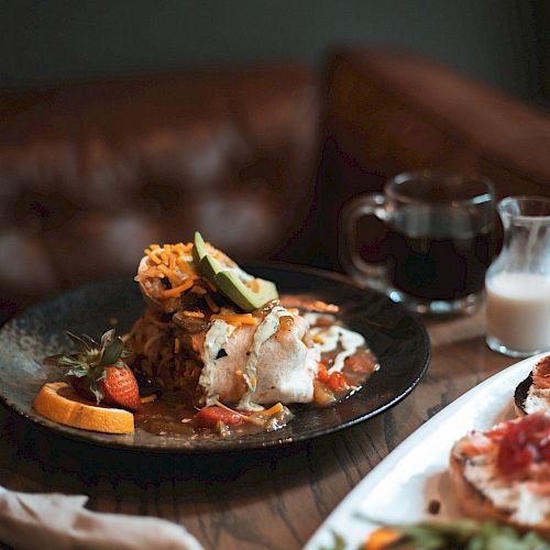 A black plate with a gourmet dish including vegetables and a small pitcher; a cup of coffee and some greens on a white plate nearby, on a wooden table.