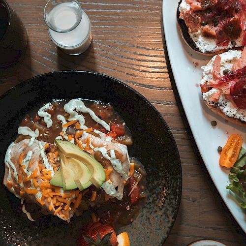 The image shows a meal with a black plate of food topped with avocado, a drink, a small glass of milk, toast with toppings, and a side salad.