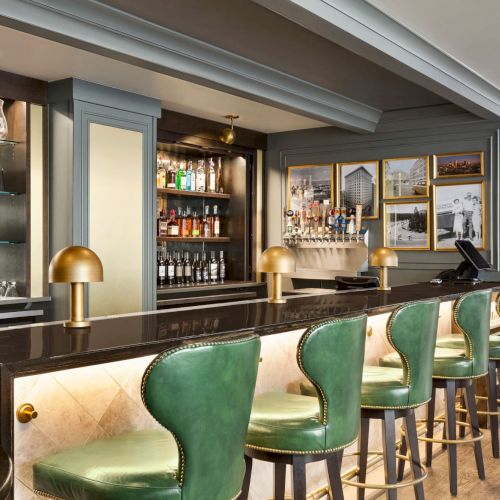 A stylish hotel bar with a stocked liquor cabinet, wine bottles, framed photos, brass pendant lights, and green upholstered stools and chairs.