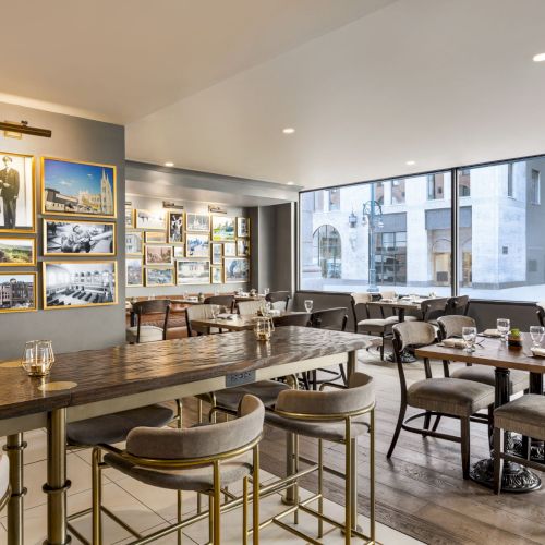 A modern dining room with a long wooden table, cushioned chairs, framed photos on the wall, and large windows along the side.