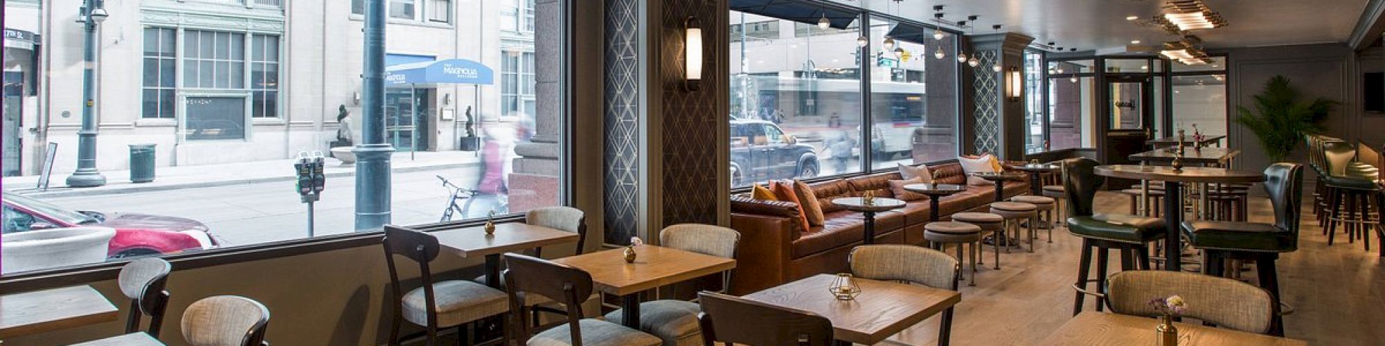 The image shows an empty, modern café with wooden tables, chairs, and large windows facing a street. Natural light fills the cozy, inviting space.