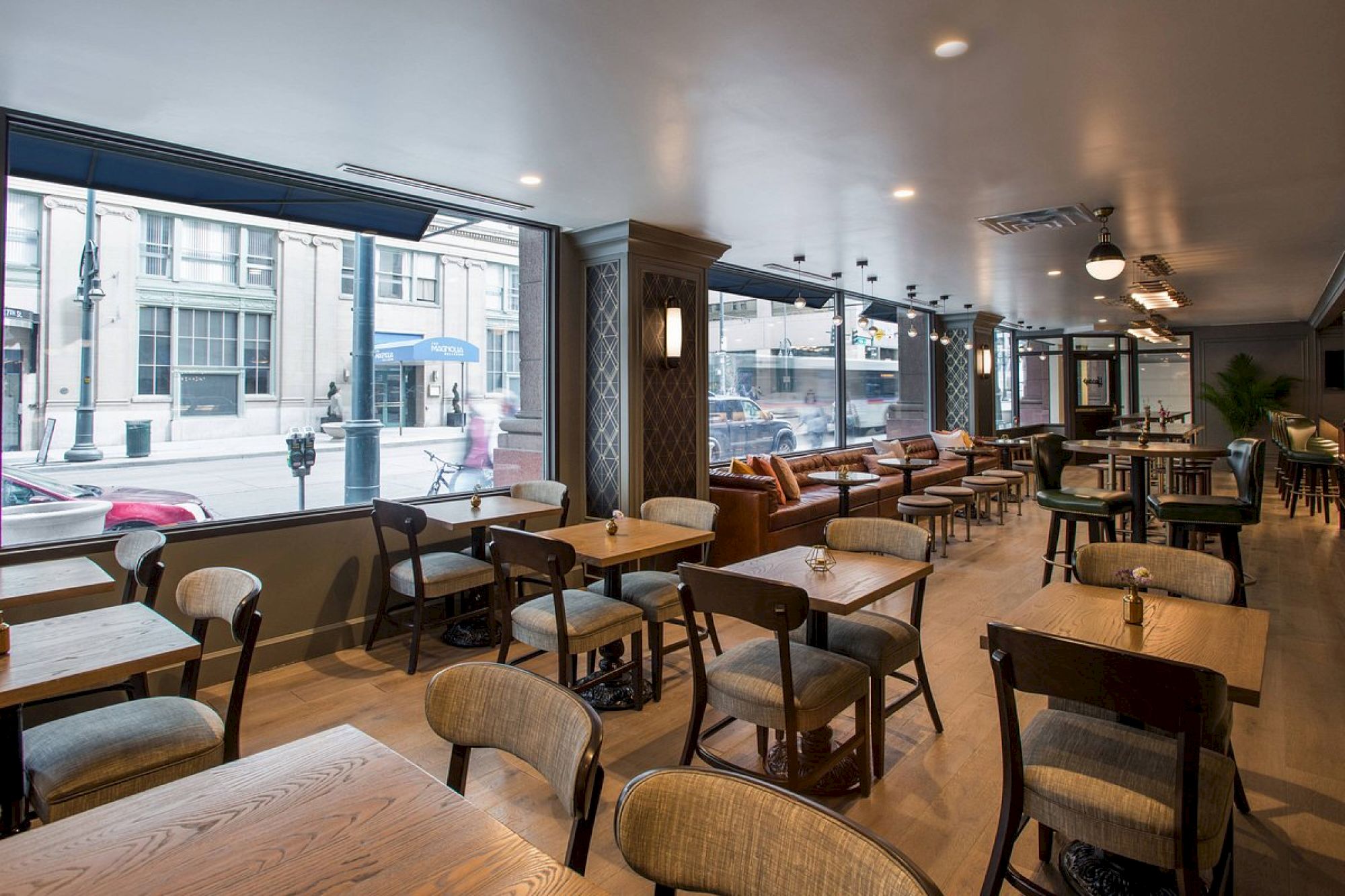 The image shows a modern, well-lit cafe or restaurant with wooden tables and chairs near large windows overlooking a city street.