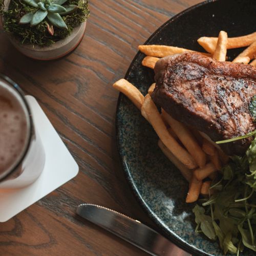 A plate with steak, fries, and greens, accompanied by a beer in a glass on a coaster and a small succulent plant.