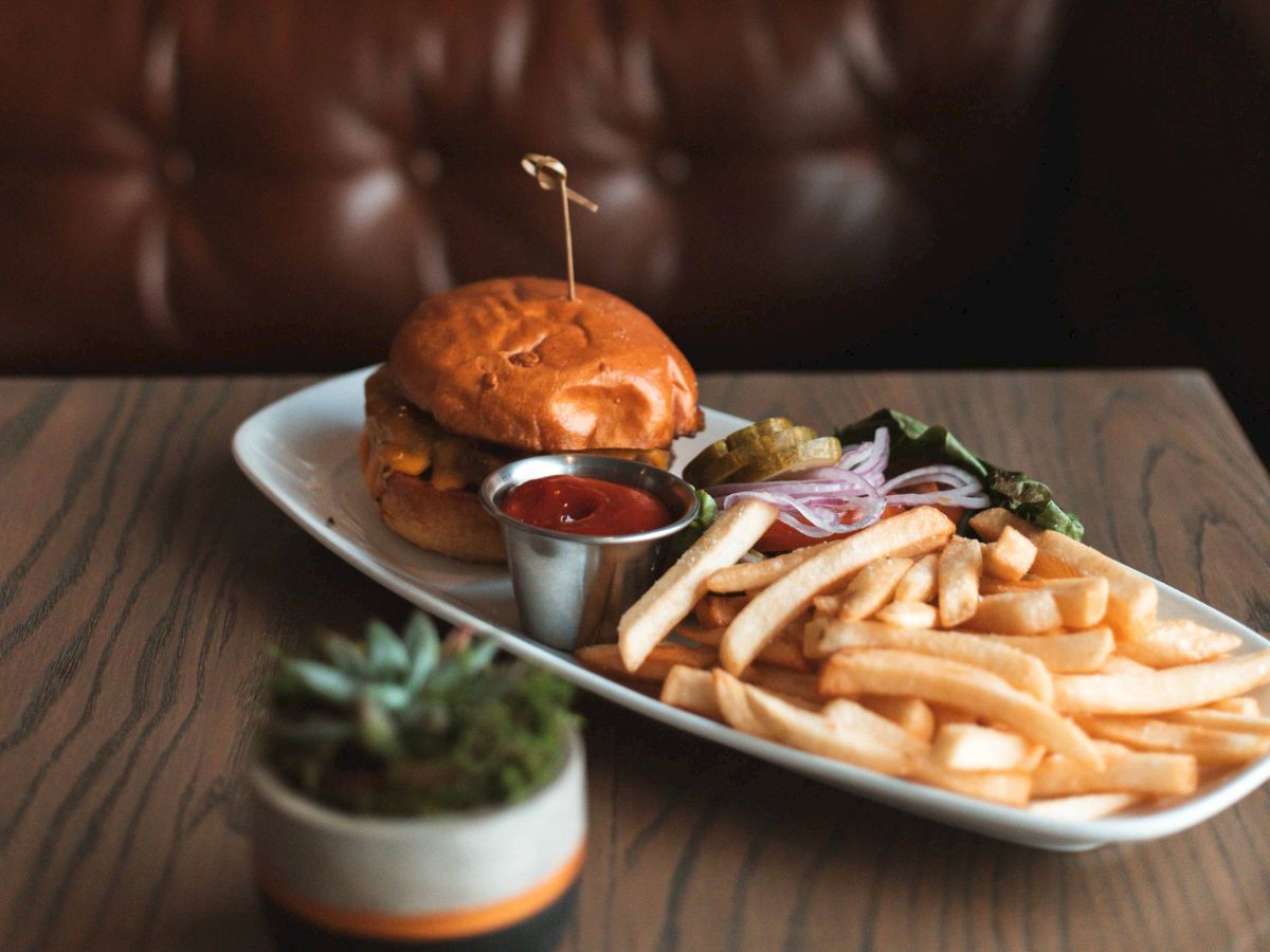 A plate with a burger, fries, ketchup, and pickles on a wooden table; a plant is in the foreground.