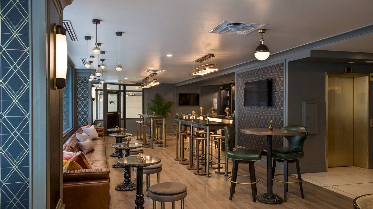 The image shows a modern, stylish lounge with a combination of high tables, stools, couches, and ambient lighting, designed for socializing.