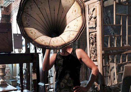 A person stands in an antique store with a phonograph horn covering their head, surrounded by vintage furniture pieces and decor.