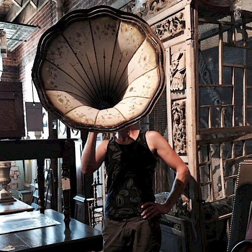 A person stands in an antique store with a phonograph horn covering their head, surrounded by vintage furniture pieces and decor.