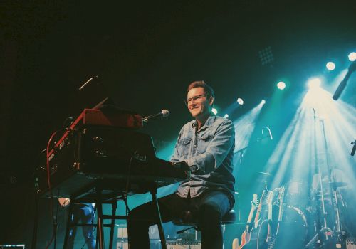 A musician playing a keyboard on stage, smiling under bright lights, with a drum set and guitars in the background.