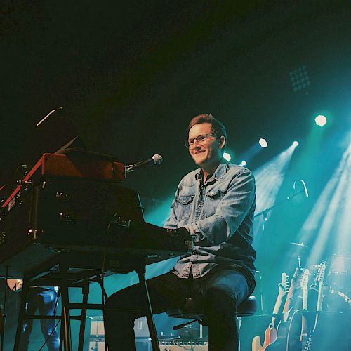 A musician playing a keyboard on stage, smiling under bright lights, with a drum set and guitars in the background.