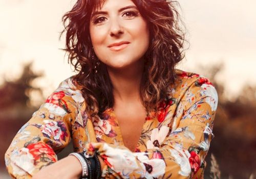 A woman with curly hair is sitting outdoors, wearing a floral blouse and bracelets, and smiling at the camera, with a blurred natural background.