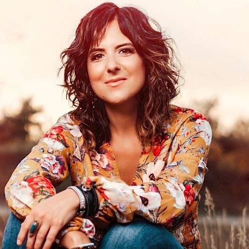 A woman with curly hair is sitting outdoors, wearing a floral blouse and bracelets, and smiling at the camera, with a blurred natural background.