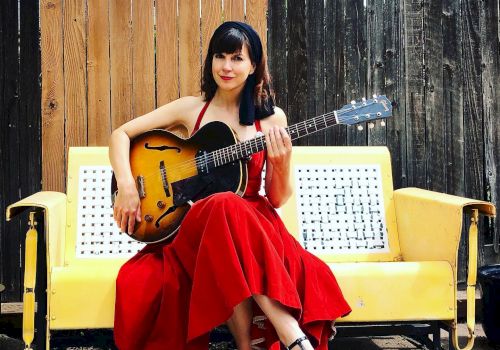 A woman in a red dress sits on a yellow bench, holding a guitar. She is outdoors with a wooden fence in the background, ending the sentence.