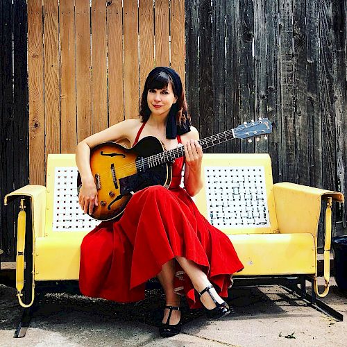 A woman in a red dress sits on a yellow bench, holding a guitar. She is outdoors with a wooden fence in the background, ending the sentence.