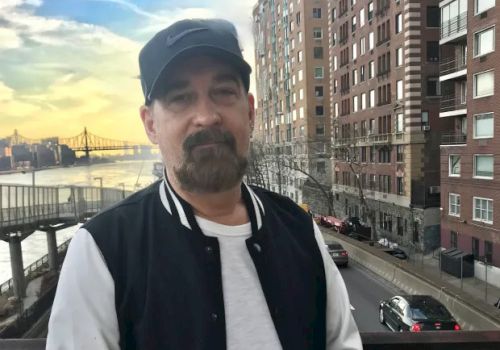 A man wearing a black cap and jacket stands in front of a cityscape with tall buildings, a bridge, and a river in the background.