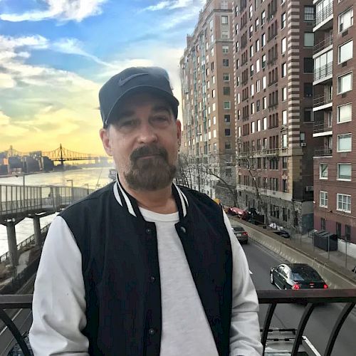 A man wearing a black cap and jacket stands in front of a cityscape with tall buildings, a bridge, and a river in the background.