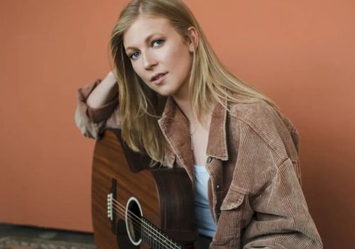 A person with long blonde hair is sitting against a peach-colored wall, wearing a brown shirt and holding an acoustic guitar, looking at the camera.